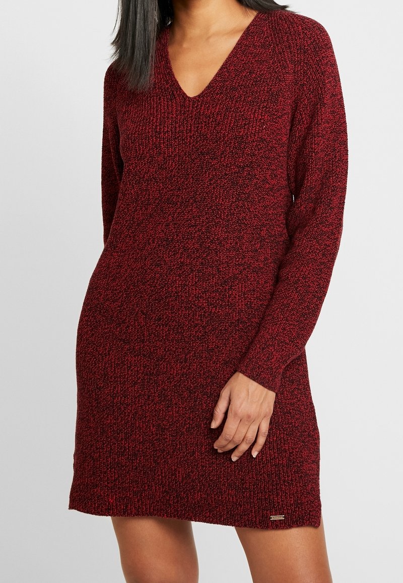 Femme portant une robe pull en maille rouge et noire à manches longues avec un col en V, debout devant un fond clair uni.