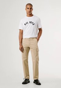 Fehér, rövid ujjú póló fekete "PJL 1973" felirattal, bézs színű, oldalzsebes cargo nadrággal és fekete cipőkkel.