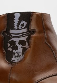 Braune Leder-Ankle-Boots mit einem schwarzen elastischen Einsatz, der ein Schädel- und Pik-Ass-Motiv zeigt. Glatte Oberfläche mit detaillierter Naht.