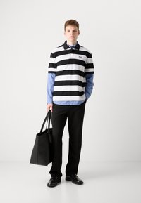 Svart-hvite stripete polo skjorte lagd over en lyseblå skjorte, paret med svarte bukser og en svart totebag. Svarte sko fullfører antrekket.