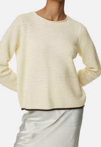 Sweter w kolorze kremowym z teksturowanym wzorem w wafelkowe sploty. Posiada okrągły dekolt oraz brązowy dół, zestawiony z gładką, jasną spódnicą.