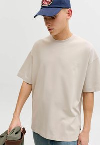 T-shirt surdimensionné beige clair en coton doux avec un col rond. Le modèle tient un vêtement vert plié, portant une casquette bleue avec un logo.