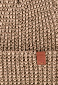 Czapka dzianinowa w kolorze jasnobrązowym z grubym, teksturowanym wzorem. Posiada brązową skórzaną metkę z wytłoczonym napisem informującym o marce.