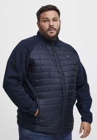 Tamsiai mėlynas zip-up tipo džemperis su pagalvotu priekiniu paviršiumi, minkštais audinio rankovėmis ir šoninėmis kišenėmis. Dizainas pabrėžia komfortą ir šilumą.