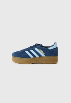 Tênis de camurça azul-marinho com riscas azul claro, biqueira redonda e sola em borracha cor de goma. Logótipo dourado "Spezial" na lateral, design clássico da Adidas.