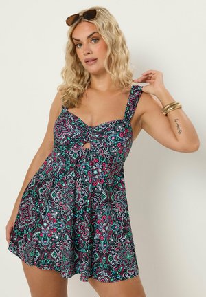 Femme blonde portant des lunettes de soleil, robe sans manches à motif floral coloré, avec des bracelets et un tatouage sur l'avant-bras.