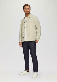 Veste boutonnée gris clair avec une poche poitrine, portée sur un t-shirt blanc, associée à un pantalon en denim foncé et des baskets blanches.