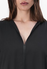 Haut noir à col en V avec fermeture éclair, fabriqué dans un tissu lisse avec un léger éclat et une coupe décontractée.