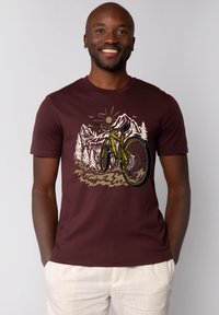 Bordeauxfarbene Baumwoll-T-Shirt mit einem Grafikdesign eines Fahrrads in grünen Akzenten, umgeben von Bergen und Bäumen, vor einem sonnenbeschienenen Hintergrund.