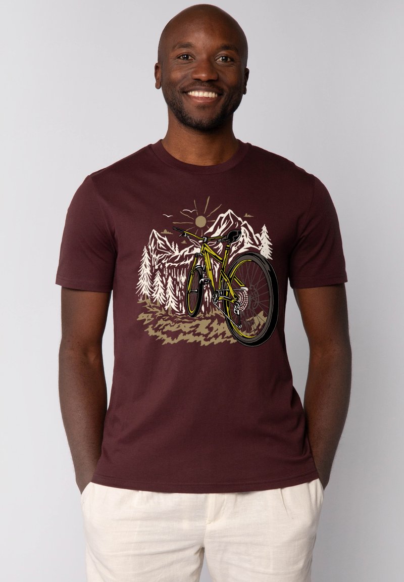 Bordeauxfarbene Baumwoll-T-Shirt mit einem Grafikdesign eines Fahrrads in grünen Akzenten, umgeben von Bergen und Bäumen, vor einem sonnenbeschienenen Hintergrund.