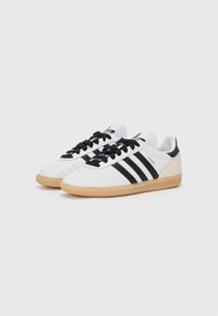 White sneakers with black stripes, textured leather and synthetic upper, round toe, black laces, and gum rubber sole. "SAMBA" logo in gold.