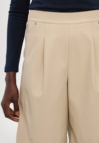 Beige plisserte bukser med en jevn tekstur, sidelommer og sølvfarget detaljer. Modellen har hånden hvilende på stoffet.
