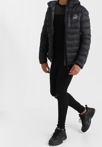 Gepolsterte schwarze Jacke mit Kapuze und einem Logo auf der Brust, getragen über einem schwarzen, strukturierten Pullover und schwarzen Slim-Fit-Jeans. Schwarze Sneaker.