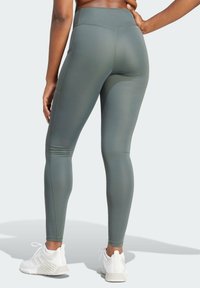 Leggings a vita alta, aderenti, in un tessuto verde tenue, caratterizzati da cuciture eleganti e una texture liscia, abbinati a scarpe da ginnastica bianche.