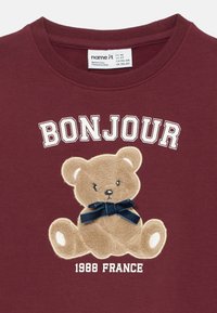 Bordo sporta krekls ar mīksta teddy bears aplikāciju un tumši zilu loku. Virs lāča ar baltu diegu izšūta vārds "BONJOUR" un "1988 FRANCE".