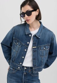 Jeans jakna tamnoplave boje s metalnim kopčama, dva džepa na prsima i skraćenim dizajnom. Nošena preko bijele majice.