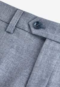 Pantalon en tissu texturé gris avec un devant plat, comportant un bouton bleu et une passante de ceinture, présentant un motif tissé subtil.