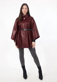 Capa de cuero sintético color burdeos con cuello alto, cintura ceñida con un cinturón negro y mangas acampanadas. Combinada con jeans ajustados gris oscuro y botas negras.