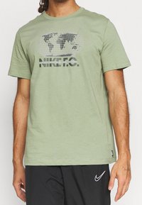 Camiseta de algodón en verde oliva con un gráfico de un globo y el texto "NIKE F.C." en blanco y negro, con cuello redondo y mangas cortas.