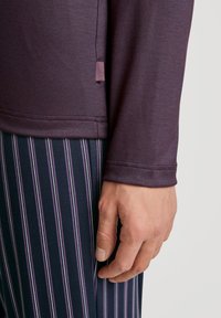Haut à manches longues violet foncé avec une texture lisse, arborant une discrète étiquette de logo. Associé à un pantalon rayé marine avec des lignes violettes et blanches.