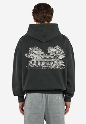 Mann, der einen schwarzen Hoodie trägt und auf dem Rücken eine weiße Skizze eines Hauses mit Bäumen und den Text "Moments to Memories" zeigt, kombiniert mit grauer Hose.
