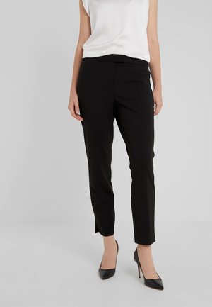 Femme portant un pantalon noir ajusté aux chevilles, un haut blanc sans manches et des chaussures à talons hauts noires, debout devant un fond gris uni.