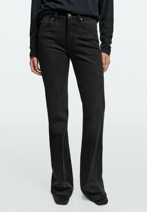 Person trägt hoch taillierte schwarze Jeans mit geradem Bein und ein langärmeliges schwarzes Oberteil vor einem einfarbigen hellen Hintergrund.