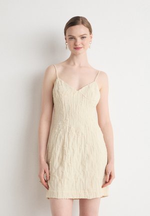 Beige kjole med tynne stropper, v-hals, teksturert stoff og en tettsittende silhuett, med subtile rynkedetaljer gjennom hele.