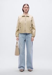 Μπεζ κοντό σακάκι με φερμουάρ μπροστά, δύο πλαϊνές τσέπες και ελαστικά μανίκια, σετάρισμένο με ανοιχτό μπλε τζιν παντελόνι φαρδύ και μπεζ τσάντα.