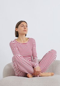 Σετ με μακρυμάνικη μπλούζα και παντελόνι με κόκκινες και λευκές ρίγες, εφαρμοστό στυλ, μαλακό ύφασμα, φορεμένο από ένα άτομο που κάθεται πάνω σε μια ελαφρώς υφασμένη επιφάνεια.