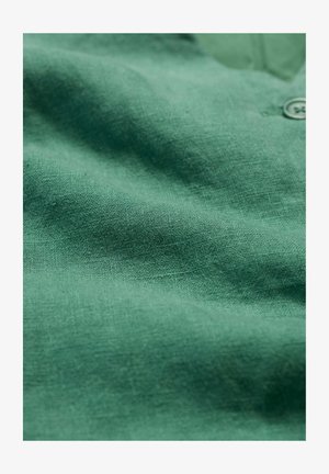 Grüner Leinenstoff mit einer strukturierten, gewebten Oberfläche. Ein Knopf ist teilweise sichtbar und kontrastiert mit dem glatten Fall des Materials.