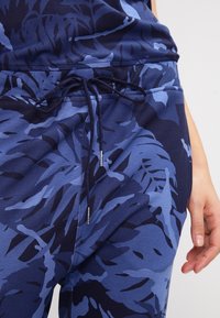 Pantaloni jogger blu navy con un grande motivo a foglia in diverse sfumature di blu, dotati di vita con cordoncino e dettagli in metallo argentato.