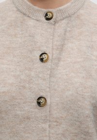 Cardigan en maille beige douce avec trois grands boutons ronds en écaille de tortue et un col rond côtelé.