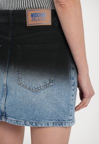 Denim svārki ar izbalēšanu no melnas uz gaiši zilu, ar ādas birku, kas marķēta "MOSCHINO JEANS", un divām aizmugurējām kabatām.