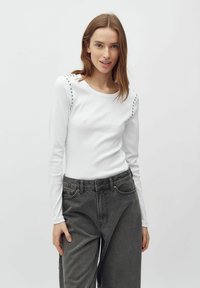Top bianco a maniche lunghe a coste con borchie silver sulle spalle, abbinato a jeans grigi scuri a vita alta. Texture del tessuto liscia.