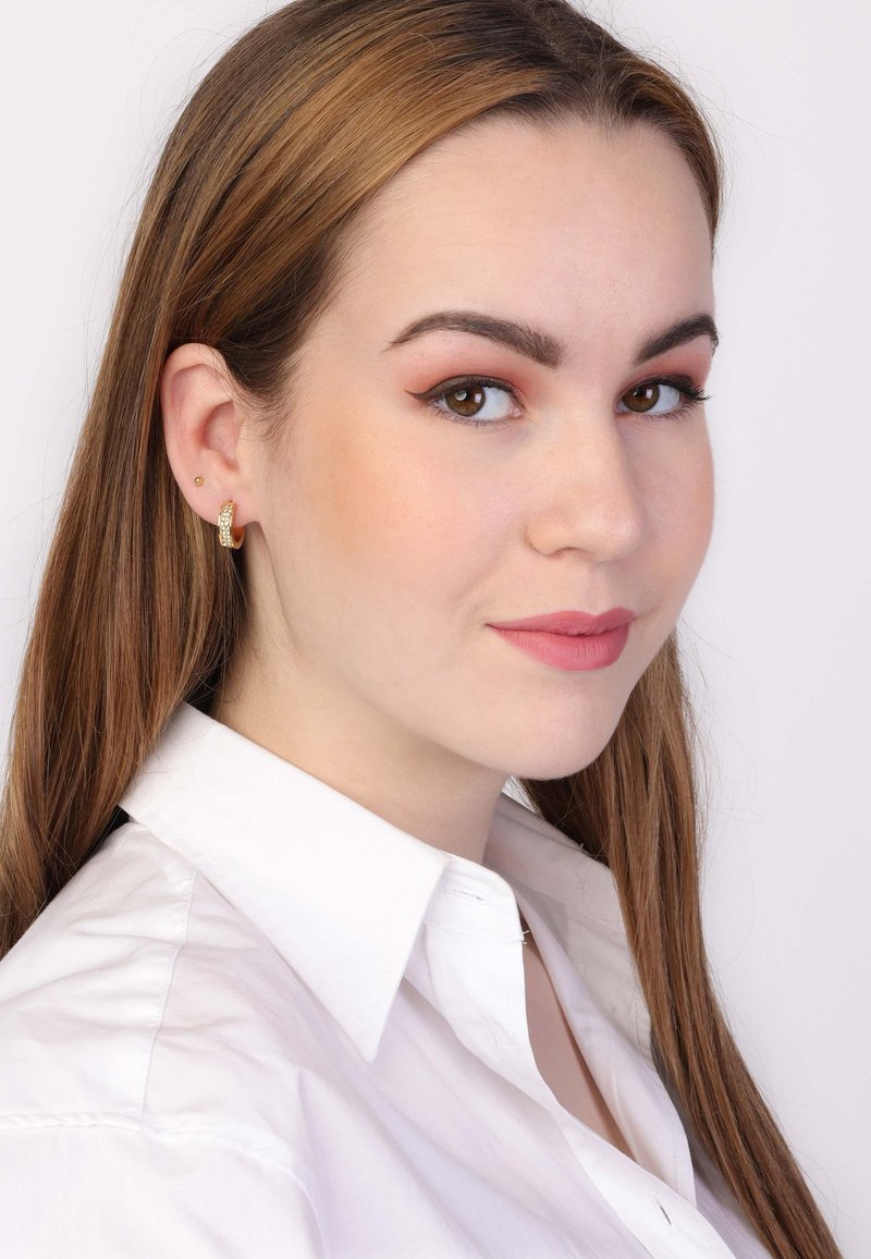 Boucles d'oreilles en or en forme de créole avec un design texturé et un petit clou dans l'oreille d'un modèle, portant une chemise blanche à col contre un fond clair.