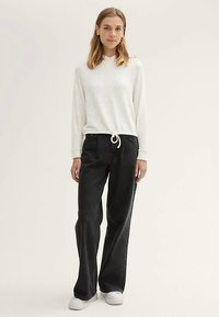 Kreminės spalvos marškiniai ilgomis rankovėmis su raišteliu juosmens srityje, derinami su plačiomis juodomis kelnėmis ir baltais sportbačiais. Paprastas, kasdieniškas aprangos stilius.