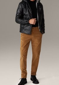 Man in een zwart leren jack, zwart shirt, bruine corduroy broek en zwarte veterschoenen tegen een neutrale achtergrond.