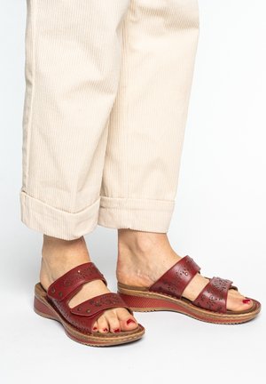 Füße in roten Leder-Wedgesandalen mit ausgeschnittenen Blumenmotiven, kombiniert mit beigen, hochgekrempelten Cordhose.