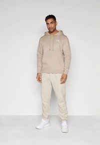 Vīrietis stāv, uzvilcis beidge Under Armour kapuci, gaišas beidge bikses un baltus sporta apavus, fona ir vienkārša balta siena.