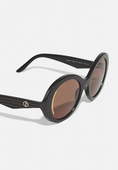 Black oversized sunglasses with round frames, brown tinted lenses, and textured black stripes on arms. Gold logo accent on left arm.