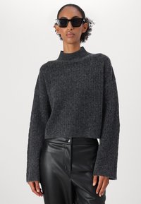 Maglione corto in maglia grigio scuro con collo alto e maniche lunghe, abbinato a pantaloni in pelle nera e occhiali da sole neri dal design angolare.