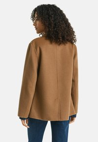Brauner Wollmantel mit lockerer Passform, der eine saubere Rückennaht und weite Ärmel zeigt, kombiniert mit blauen Jeans und einem kontrastierenden Bündchen.