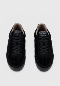 Schwarze Wildleder-Sneaker mit abgerundeter Zehenpartie, flachen schwarzen Schnürsenkeln und einer strukturierten schwarzen Gummisohle. Gestickte Details entlang der Seiten.