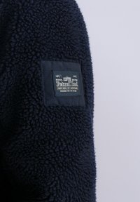 Navy blue fleece jacket, featuring a textured fabric, with a square black patch label displaying brand name and design details.