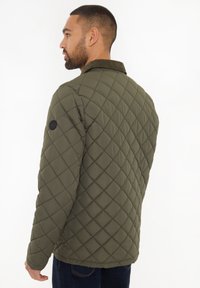Vatteret olivengrøn jakke med en kordfløjlskrave og en rund sort logo-patch på venstre ærme. Stoffet har en glat tekstur.