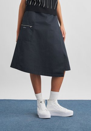 Άτομο που φοράει μπλε σκούρα φούστα μέχρι το γόνατο με τσεπάκι στο πλάι με φερμουάρ, λευκά ψηλά αθλητικά παπούτσια και λευκές κάλτσες πλήρους μήκους σε μπλε χαλί.