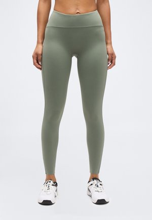 Widok z przodu osoby ubranej w oliwkowozielone legginsy z wysokim stanem oraz białe sportowe buty, stojącej na tle jednolitego jasnego tła.