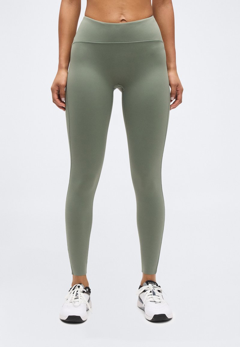 Μετωπική όψη ατόμου που φοράει ελαστικό κολάν σε χακί χρώμα με ψηλή μέση και λευκά αθλητικά παπούτσια, στέκεται μπροστά σε απλό ανοιχτό φόντο.