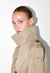 Młoda osoba z gładko zaczesanymi włosami i piegami, ubrana w beżowy trencz z podniesionym kołnierzem, oglądająca się przez ramię na tle białym.